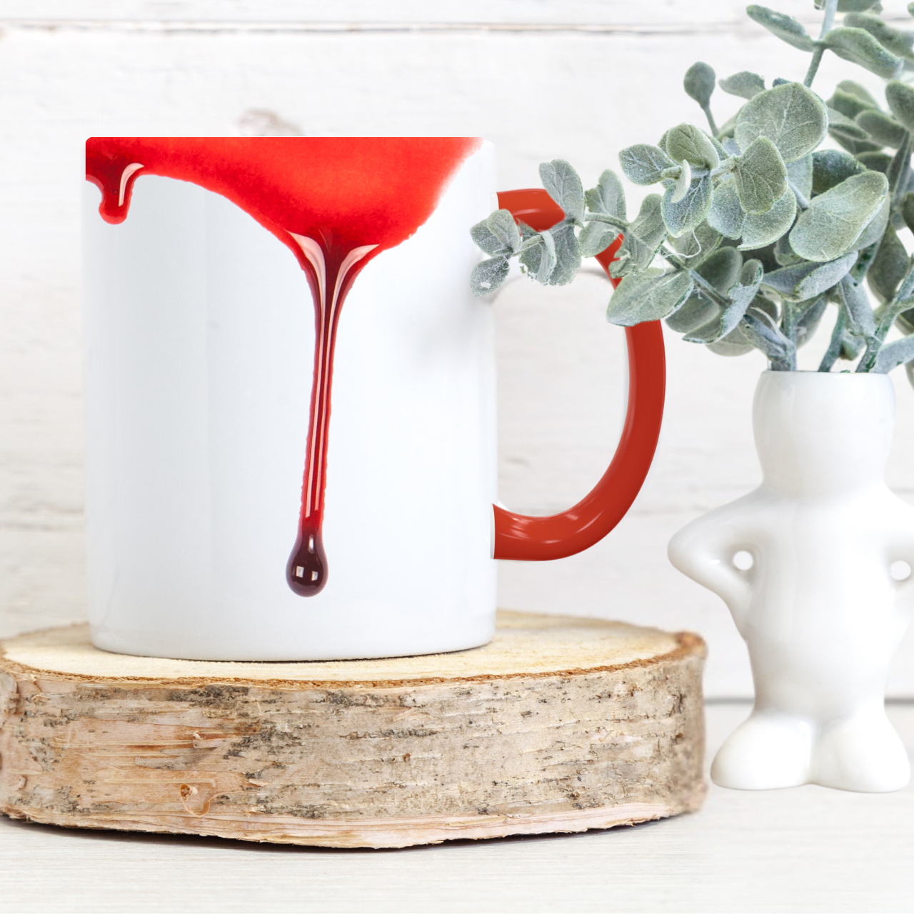 White blood dripping mug with red interior and handle on a wooden coaster, next to a plant.
