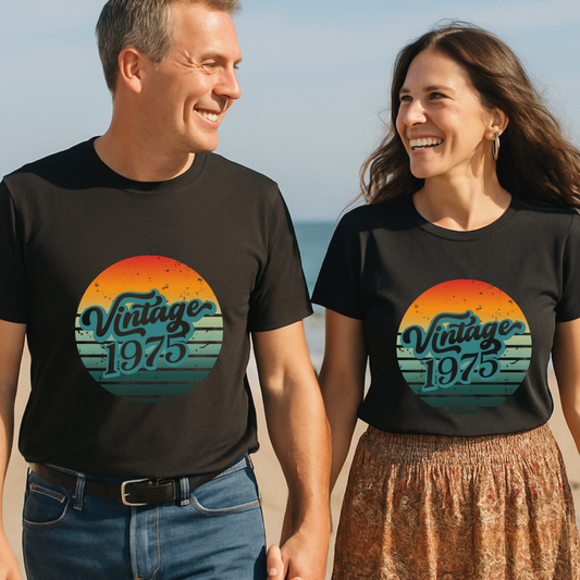 a couple in their 50s walking hand in hand along the beach waering black tshirts with the graphic of a retro sun and the words vintage 1975