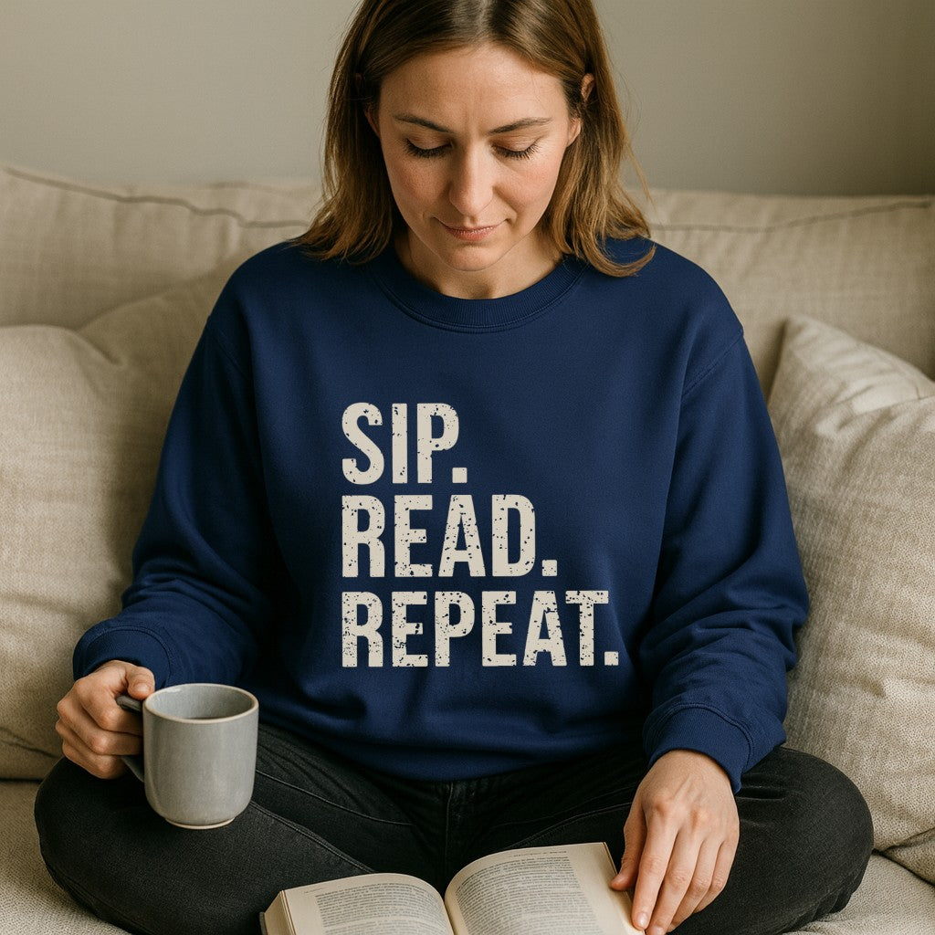 Woman wearing a navy blue sweatshirt with 'SIP. READ. REPEAT.' text, holding a mug and reading a book on a beige couch.