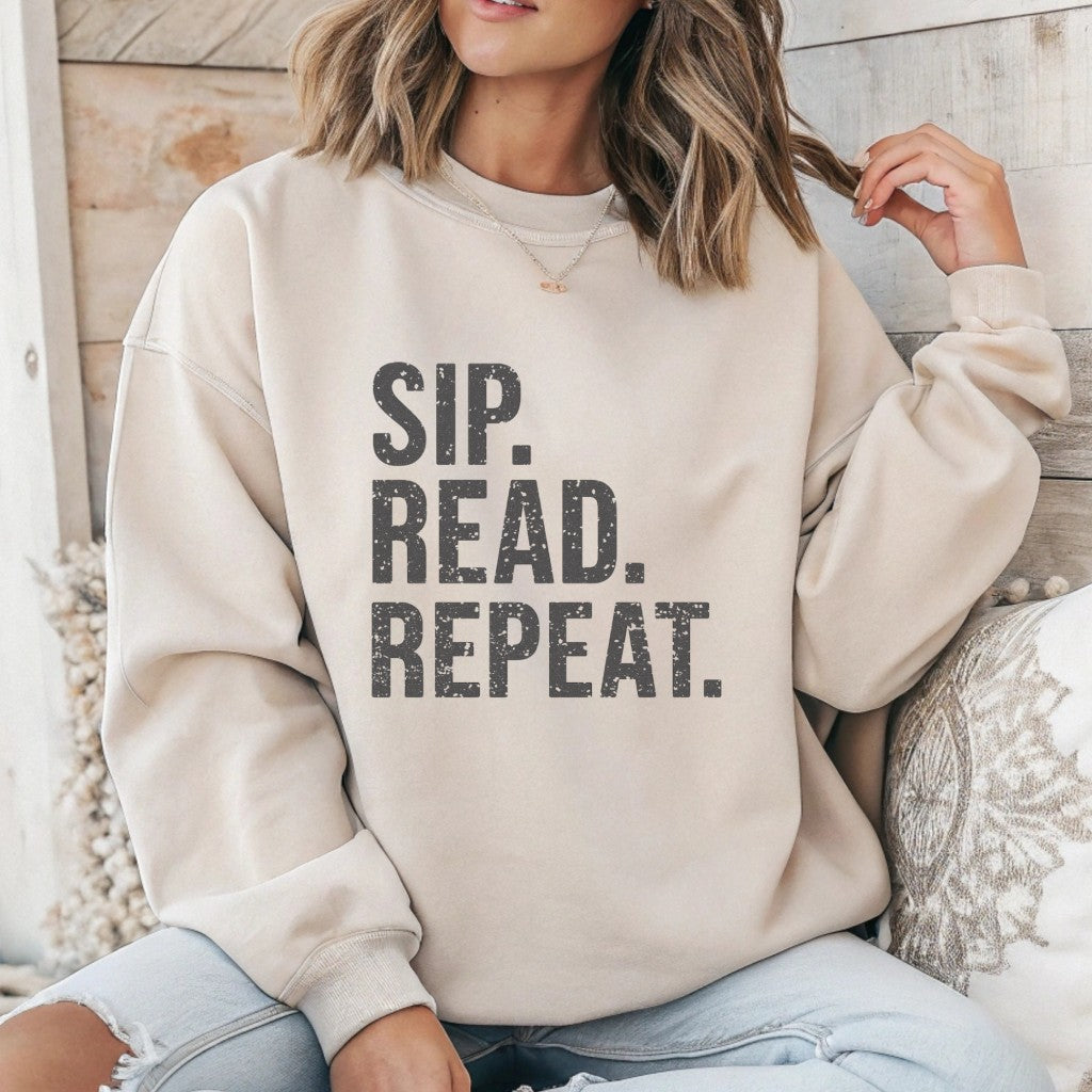 Person wearing a beige sweatshirt with 'Sip. Read. Repeat.' text, sitting against a wooden wall.