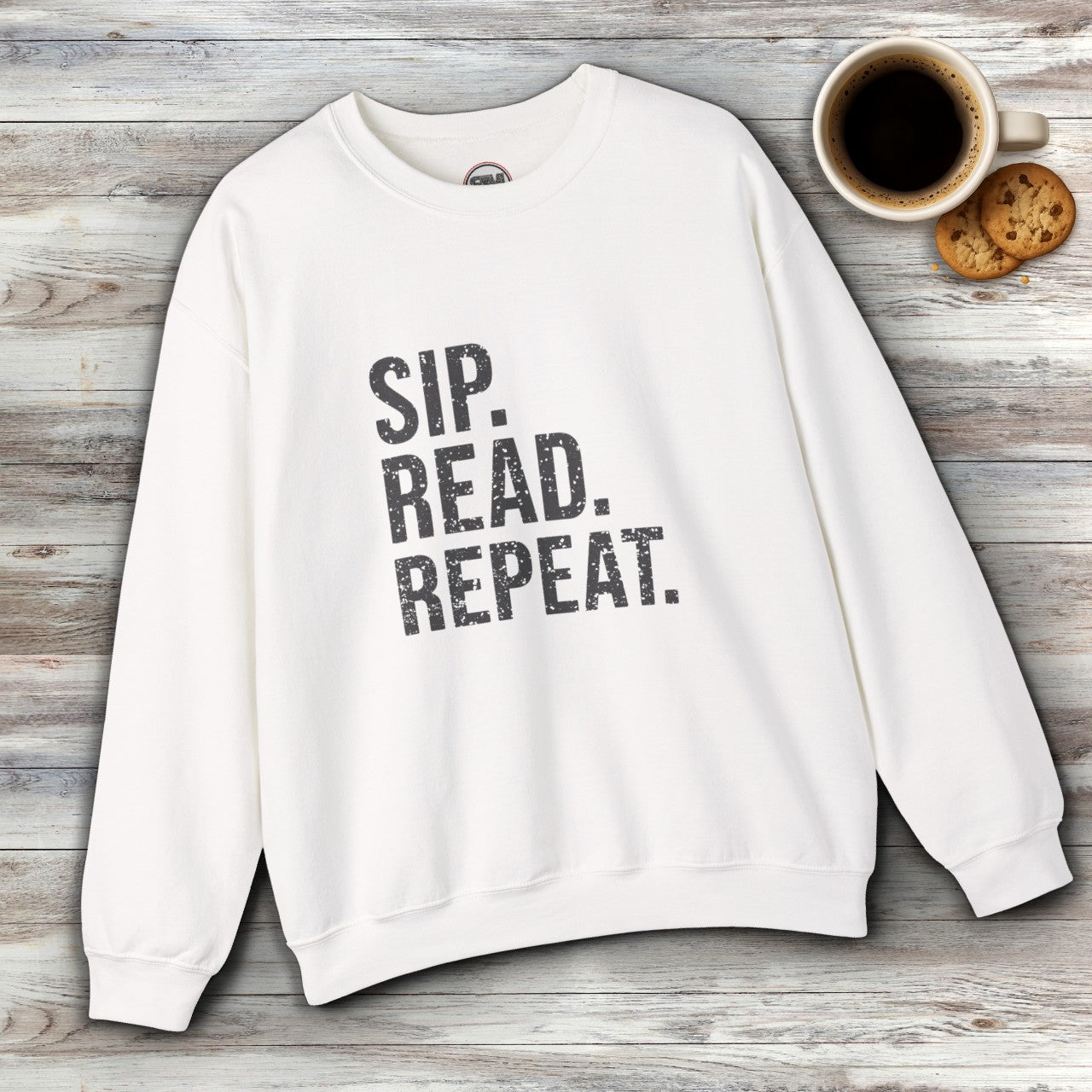White sweatshirt with 'Sip. Read. Repeat.' text on a wooden surface with coffee and cookies.