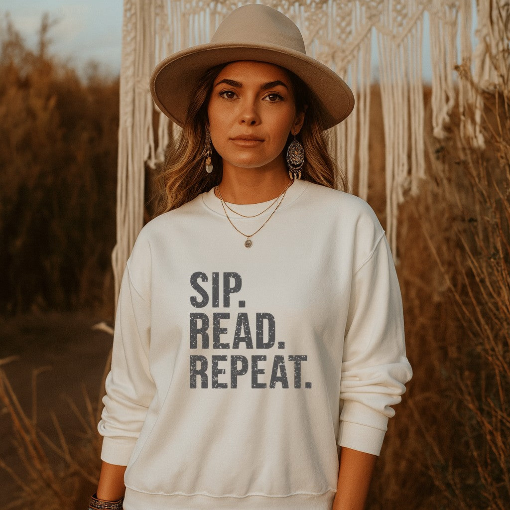 Woman wearing a white sweatshirt with 'SIP. READ. REPEAT.' text, standing in front of macramé wall.